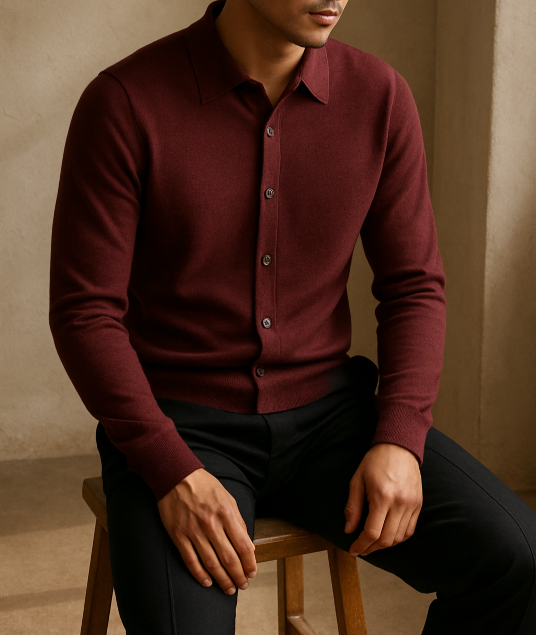 Man wearing a burgundy button-up shirt sitting on a wooden stool against a beige wall.