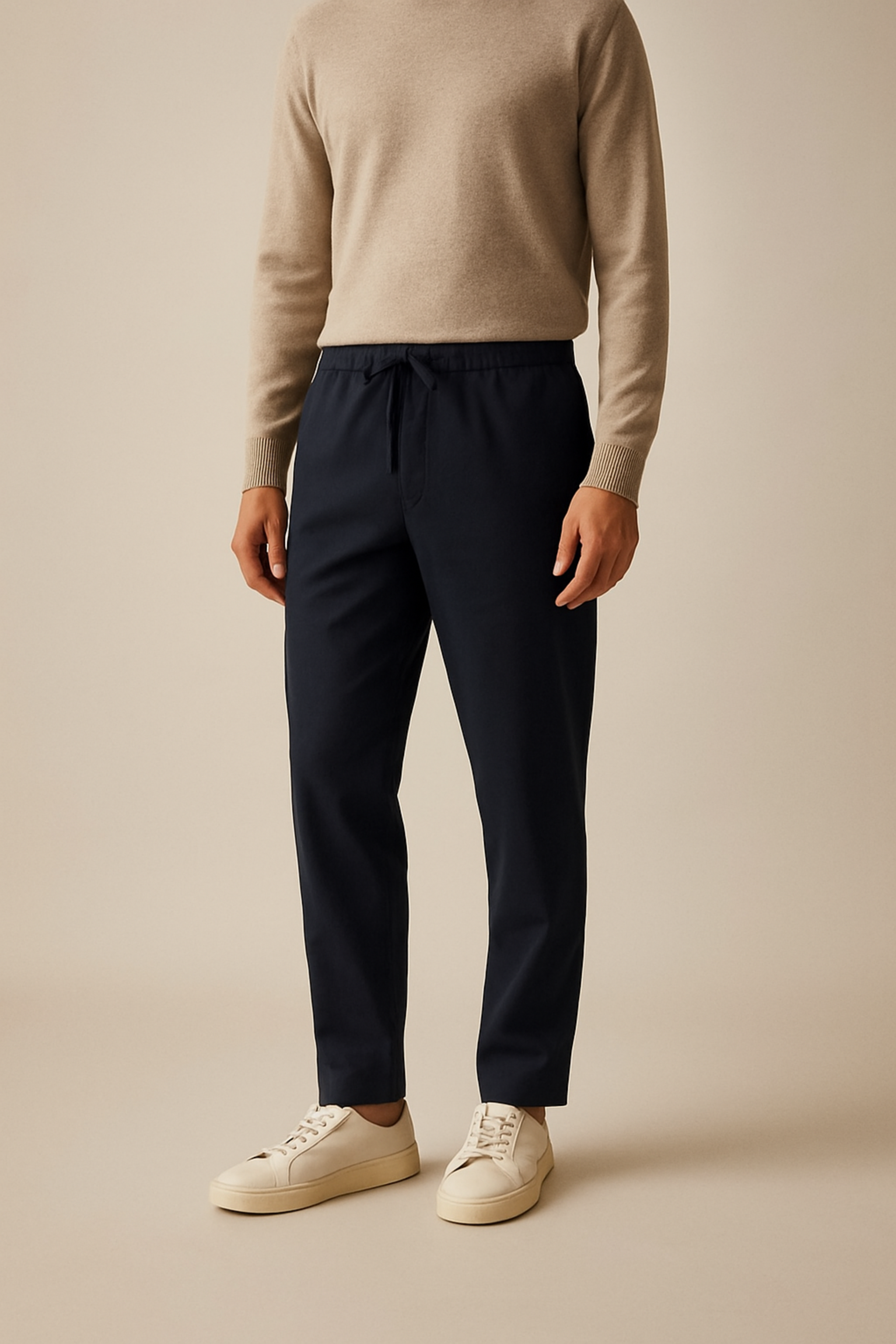 Person wearing a beige sweater and navy pants with white sneakers on a beige background